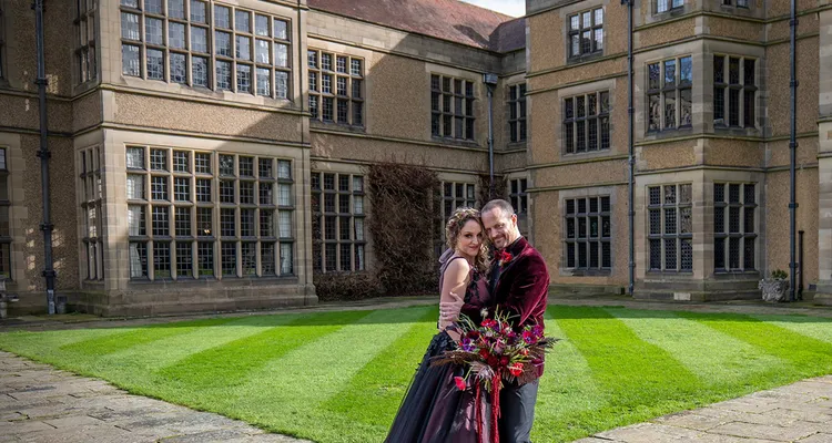Bride and groom in gothic red and black wedding outfits stand outside Fanhams Hall, a UK wedding venue