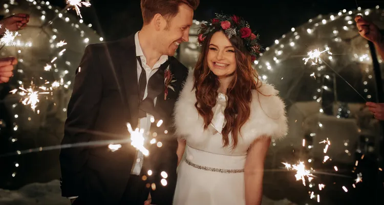 Bride and groom holding hands, with big smiles on their faces during their sparklers exit. Bride is wearing a flower headband of green and red, a white fur shawl and a white wedding dress, and the groom is wearing a dark suit with an undone bowtie