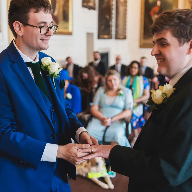 Two grooms at their wedding ceremony exchanging rings with their loved ones looking on behind them.