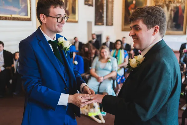 Two grooms at their wedding ceremony exchanging rings with their loved ones looking on behind them.