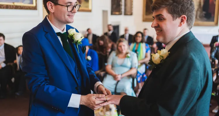 Two grooms at their wedding ceremony exchanging rings with their loved ones looking on behind them.