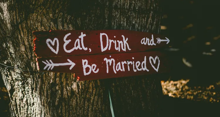 A handwritten sign saying 'eat, drink & be married' with hearts and arrows, attached to a tree