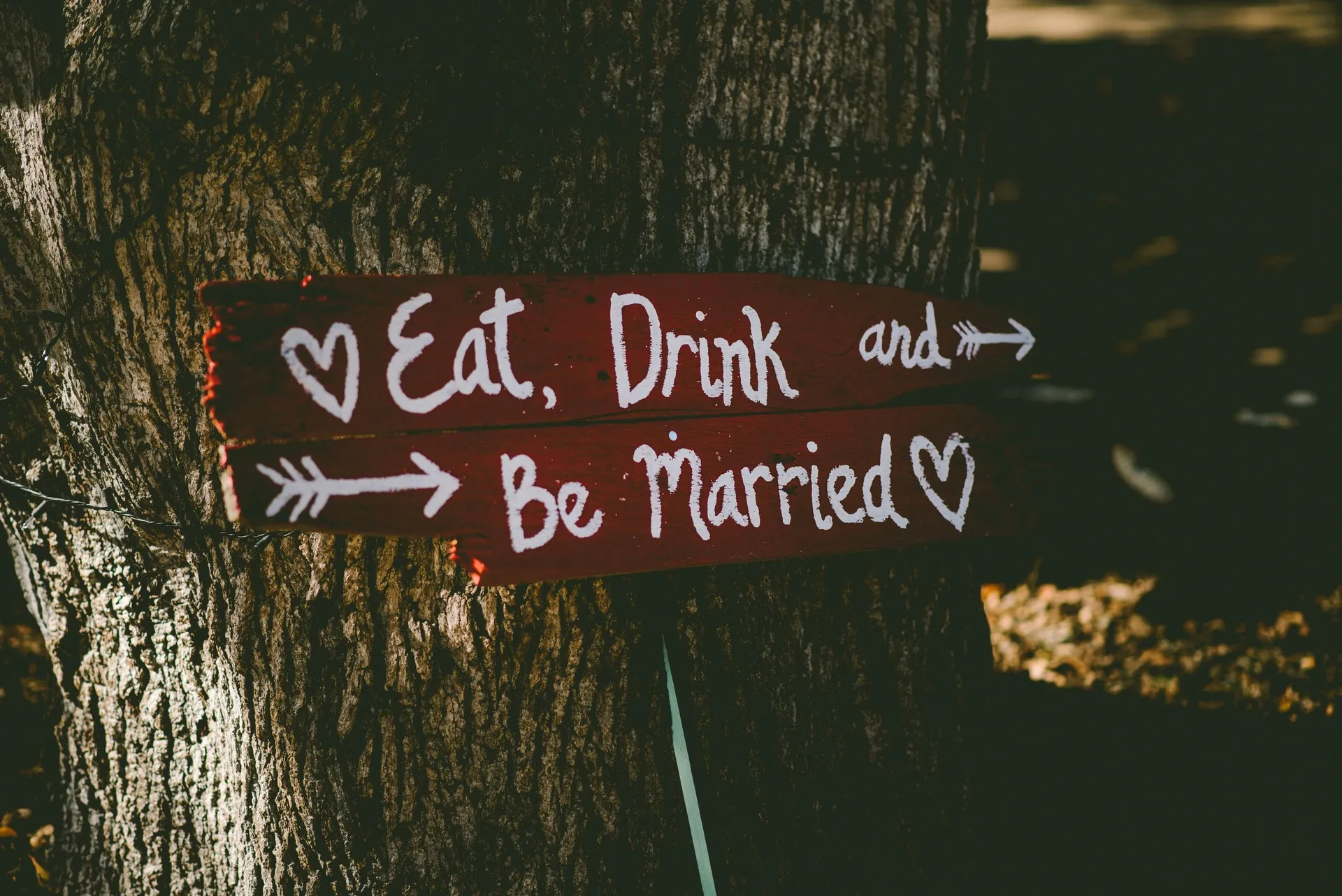A handwritten sign saying 'eat, drink & be married' with hearts and arrows, attached to a tree