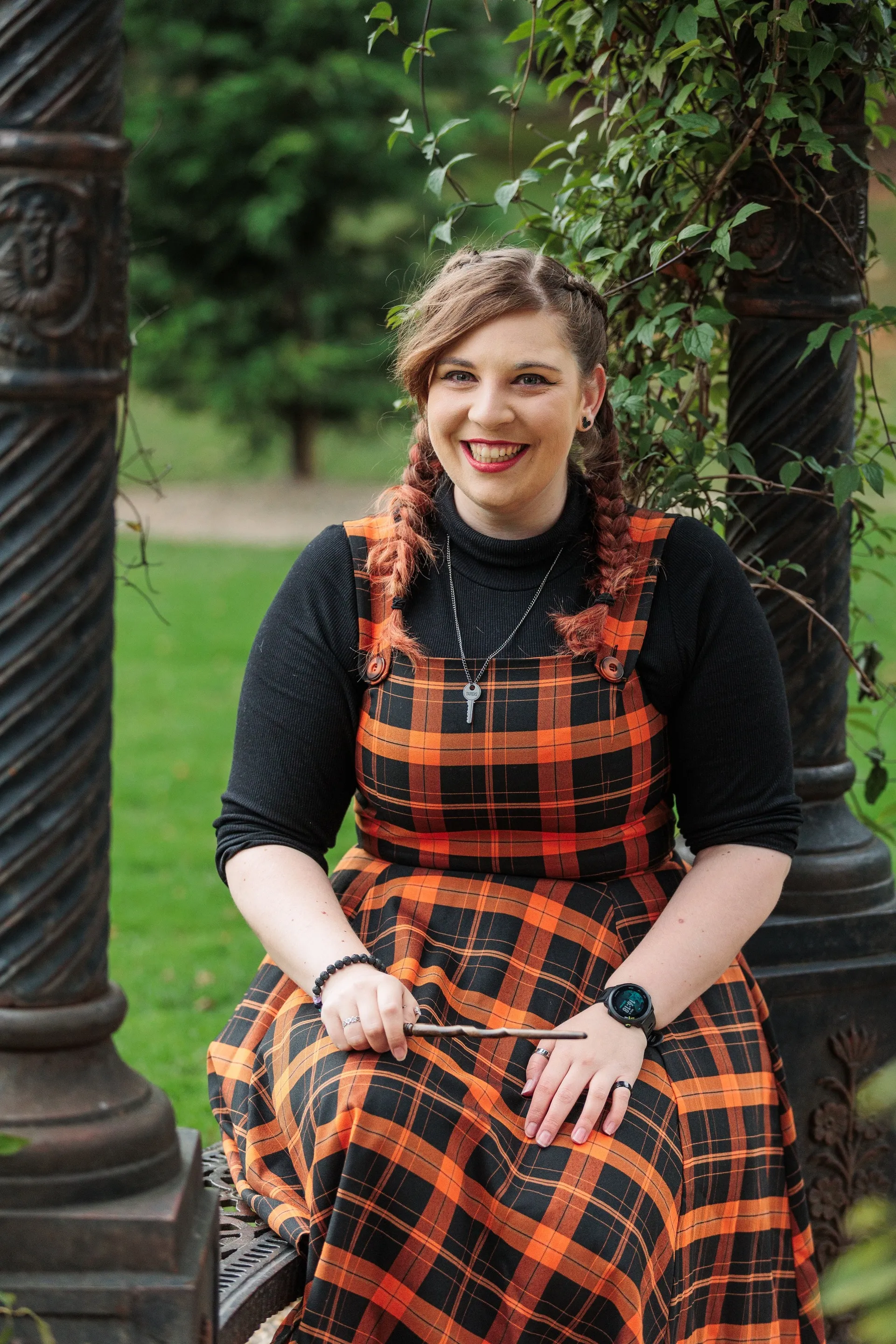 Rachel smiling to the camera sat under a metal structure with vines growing around the metal pillars. Rachel is wearing an orange, tartan dress and holding a wand.