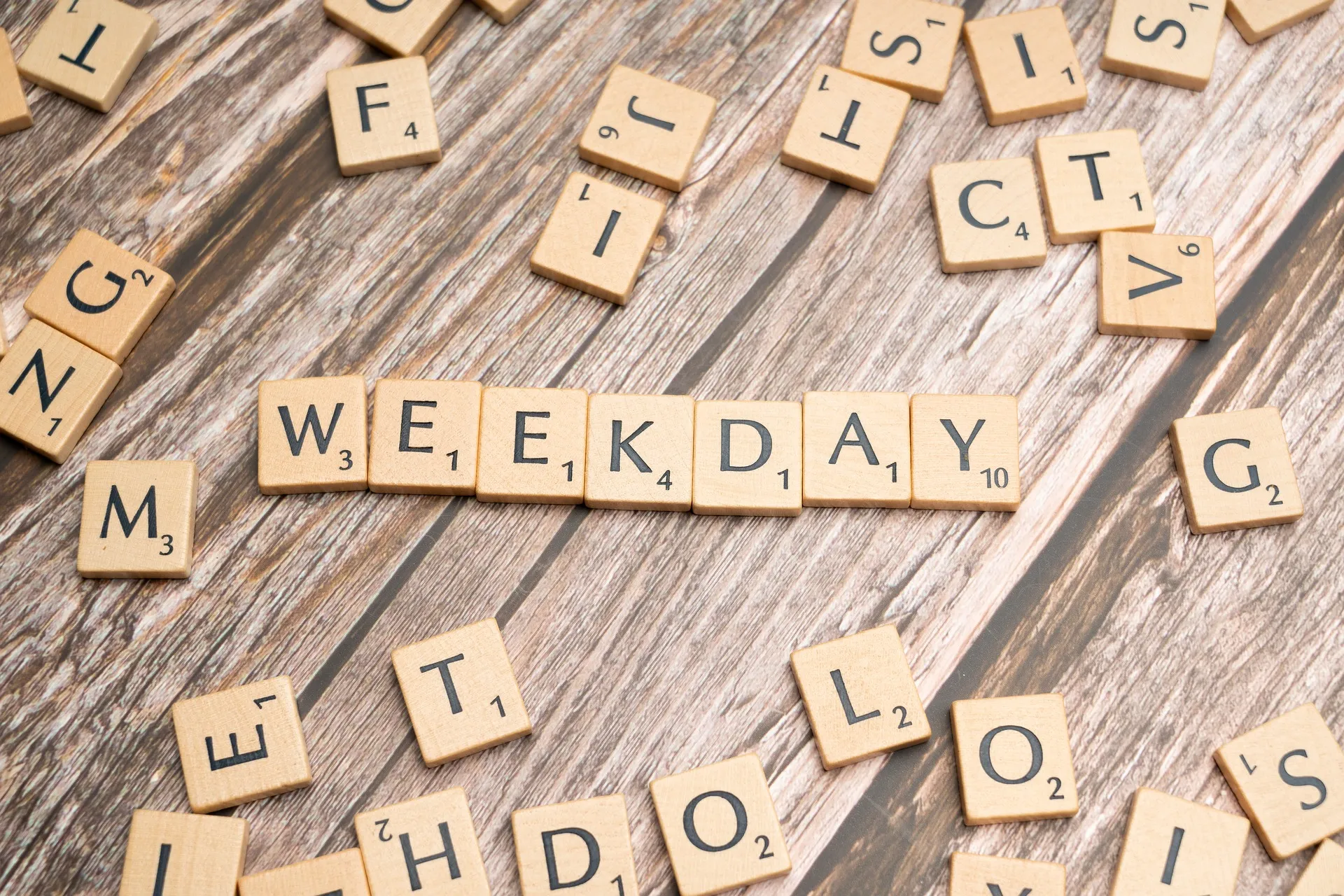 Scrabble pieces scattered on a wooden surface. At the center of the image the pieces spell out weekday