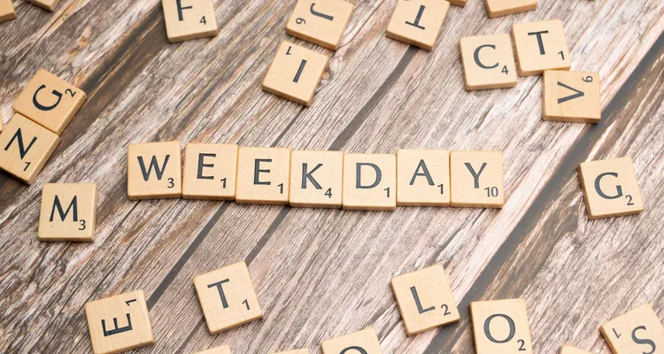 Scrabble pieces scattered on a wooden surface. At the center of the image the pieces spell out weekday