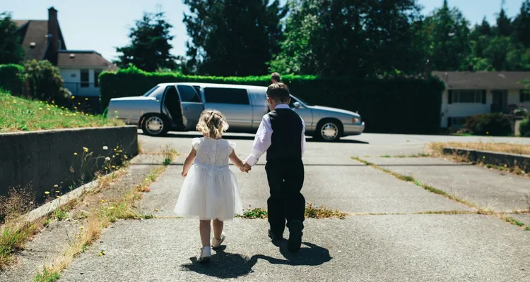 a boy in a shirt, waistcoat and black trousers and a girl in a flower girl style dress walking hand in hand on grey road during daytime towards a parked wedding car