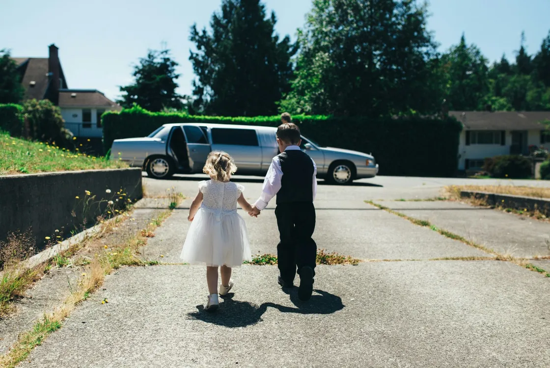 a boy in a shirt, waistcoat and black trousers and a girl in a flower girl style dress walking hand in hand on grey road during daytime towards a parked wedding car