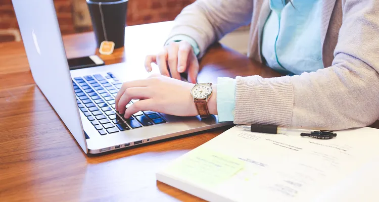 A person sat at a table ready to plan. A notebook and pen at the side, laptop open, phone close by and a hot drink