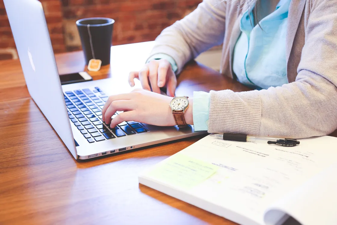 A person sat at a table ready to plan. A notebook and pen at the side, laptop open, phone close by and a hot drink